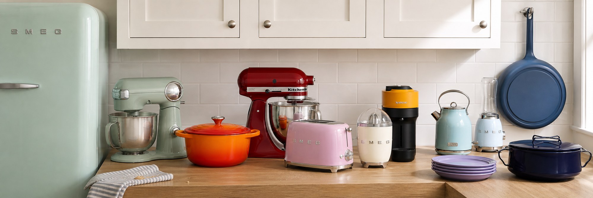 A kitchen counter set with colorful appliances: a pastel green Smeg fridge, a mint Cuisinart stand mixer, a red KitchenAid stand mixer, an orange Le Creuset casserole, a pink Smeg toaster, a yellow Nespresso machine, a turquoise Haden kettle, a pale blue Smeg blender, lavender plates and a navy pot